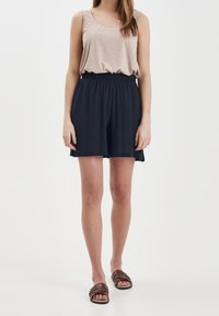 Beige ärmlös topp med rundad halsringning, kombinerad med mörkblå shorts med elastisk midjeresår. Modellen bär bruna sandaler med korsade remmar.