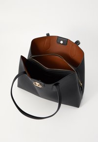 Geantă de mână din piele neagră cu un exterior neted și un interior maro. Prezintă accesorii din aur, un compartiment central cu fermoar și două mânere rezistente.