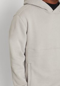 Hellgrauer Hoodie aus weichem, dickem Stoff. Verfügt über eine Kängurutasche, eine verstellbare Kapuze mit Kordelzug und sichtbare Nähte.