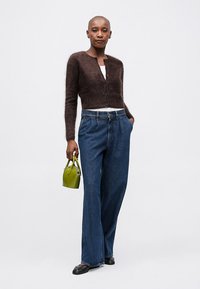 Pruun pehme kardigan valge topi peal, koos laia säärega siniste denimitega. Käes on väike roheline käekott, jalas mustad korterkingad.