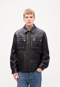 Black leather jacket with a zipper, two patch pockets with snap closures, wide collar, and a boxy fit. Worn with light blue jeans.