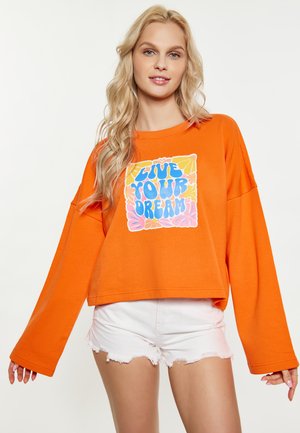 Sweatshirt orange surdimensionné avec des manches longues, présentant un graphique coloré "VIVEZ VOTRE RÊVE" sur le devant. Associé à un short blanc déchiré.