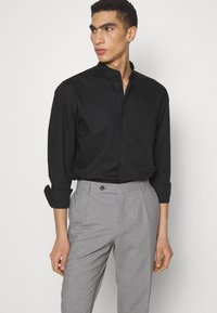 Camicia nera con colletto alla cinese, abbinata a pantaloni grigi su misura con un delicato motivo a quadri e passanti per la cintura.