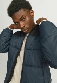 Navy blue puffer jacket with a black shoulder panel, zipper closure, and a smooth texture. Worn over a light-colored top.