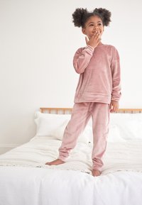 Velvet pink loungewear set featuring a long-sleeve top and slim-fit pants, worn by a child standing on a bed. Smooth texture, casual design.