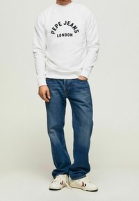 Bílý svetr s nápisem "PEPE JEANS LONDON" vytištěným černou barvou, doplněný modrými džínami a bílými teniskami s červenými a námořnickými akcenty.