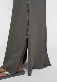 Jupe maxi en tissu vert olive avec une fente sur le côté, surface texturée et passepoil en satin noir le long de la fente, associée à des tongs argentées.