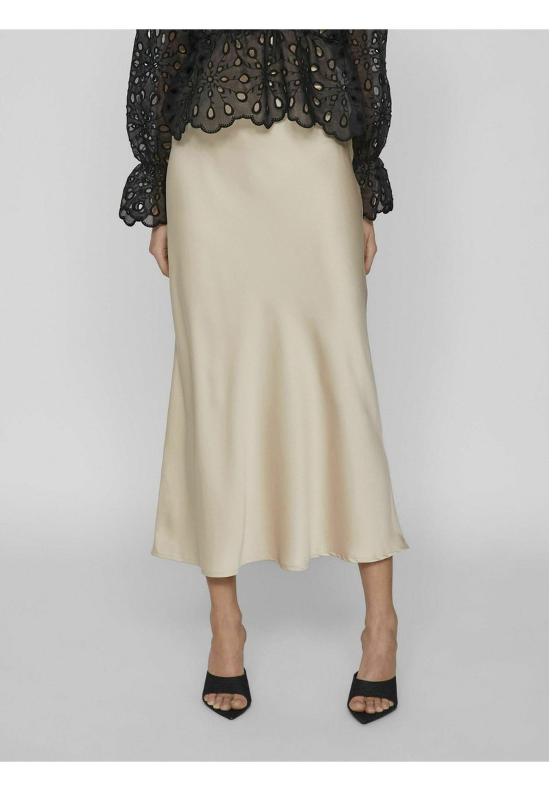Satinbeige glidkjol med en flödande design, i kombination med en genomskinlig svart topp med blommiga uttagningar och scallopade ärmar. Svarta klackmules.
