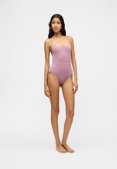 Jeune femme debout pieds nus portant un maillot de bain une pièce violet clair avec des fines bretelles sur un fond uni.