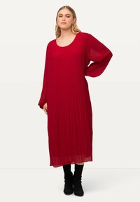 Rotes plissiertes Kleid mit langen Ärmeln, Rundhalsausschnitt und fließender Silhouette; kombiniert mit kniehohen schwarzen Stiefeln. Glatte Textur.