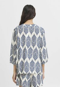 Blouse blanche avec motif floral orné bleu, manches trois-quarts et encolure ronde. Tissu doux et texturé qui tombe de manière fluide.