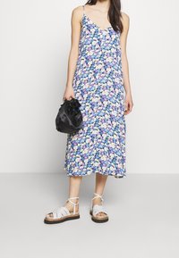 Kwiatowa sukienka midi w niebieskie, fioletowe i białe kwiaty; regulowane ramiączka spaghetti; noszona z białymi sandałkami na paski i czarną torbą.