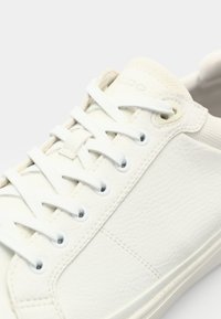 Baskets blanches en cuir texturé avec des lacets blancs, semelle en caoutchouc et petit logo près de la languette. Présente des œillets ronds.