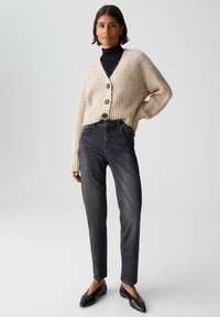 Donna che indossa un cardigan spesso beige sopra un dolcevita nero, jeans grigio scuro e ballerine nere a punta, in piedi con la mano sulla vita.