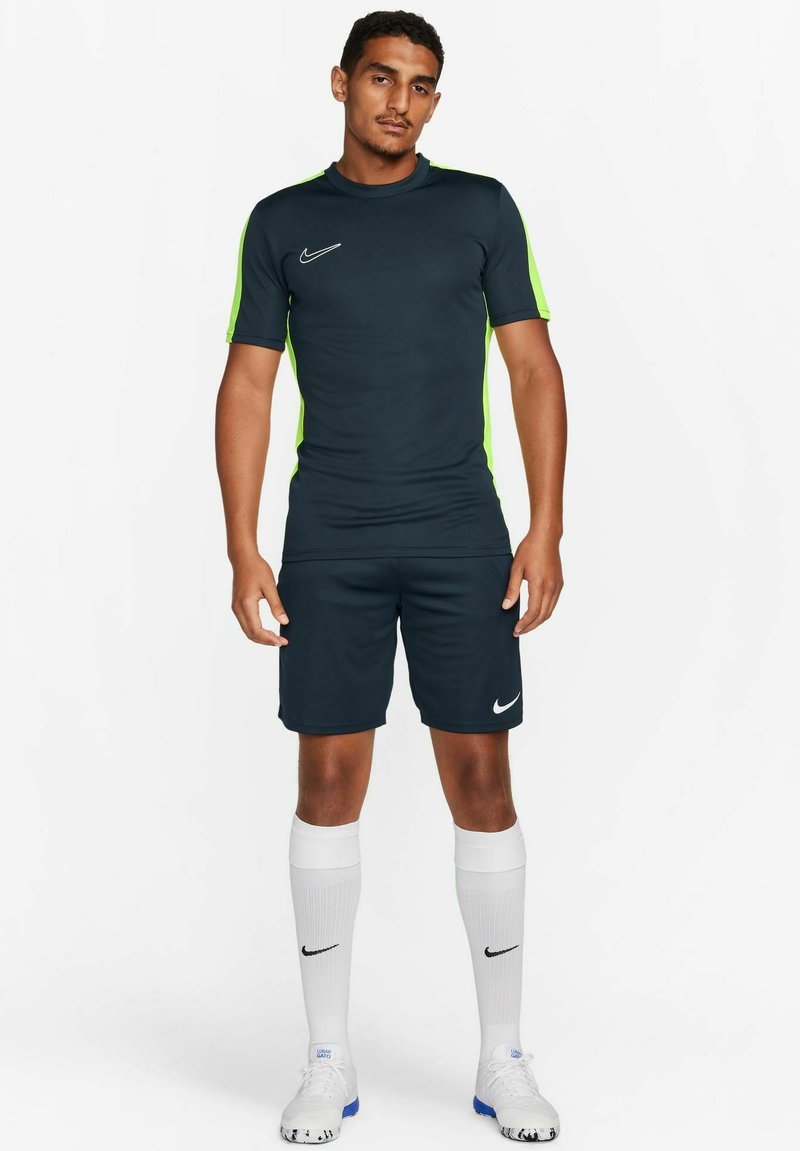 Donkerblauwe sporttrui en -short met groene accentstrepen. Bevat het Nike-logo op de borst en op de shorts. Witte kniekousen en sportschoenen.