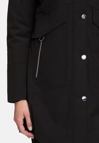 Schwarze wasserdichte Jacke mit Druckknopf- und Reißverschluss, großen Fronttaschen und glatter Textur. Akzente sind silberne Hardware-Details.