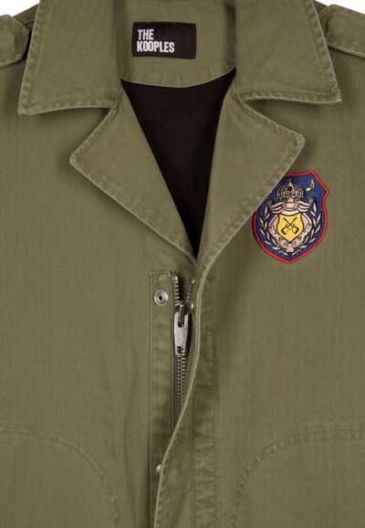 Olijfgroene jas met visgraattextuur, ritsluiting en een geborduurd embleem op de linkerborst. Label leest "THE KOOPLES."