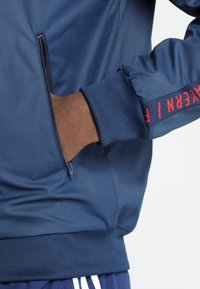 Blue bomber jacket with zippered pocket and ribbed cuffs. Features red embroidered text along the sleeve. Smooth, lightweight material.