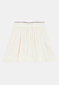 Kermakeltainen plissertu hame, jossa on takavetoketju ja hillitty brodeerattu brändäys helman lähellä.