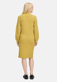Abito a maglia a maniche lunghe, giallo senape, con una silhouette aderente, polsini a coste e decorative chiusure a bottone vicino ai polsi.