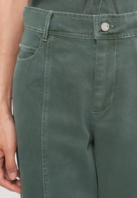 Pantaloni din denim verzi, cu un design ajustat, detalii de cusătură vizibile și un buzunar frontal cu închidere cu buton metalic.
