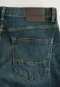 Mørkeblå denim jeans med en struktureret finish. Indeholder et brunt lædermærke med teksten "NEXT DENIM SUPPLY" og broderede detaljer på baglommen.
