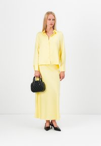 Yellow button-up shirt with long sleeves paired with a matching yellow skirt. Black, textured handbag held in right hand, and black pointed heels.