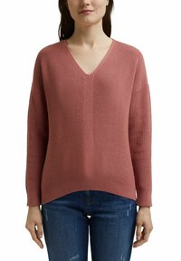 Pull à col en V, ample, en maille texturée corail douce. Manches côtelées et ourlet, associé à un jean bleu déchiré.