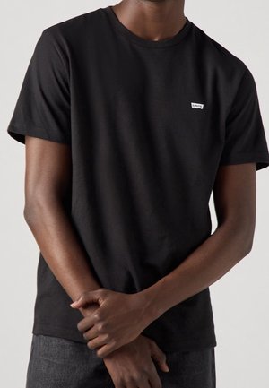 Homme portant un T-shirt Levi's noir uni à manches courtes avec un petit logo blanc sur la poitrine, les mains jointes devant, jean foncé visible.