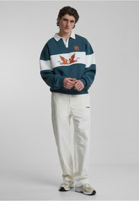 Tricou de rugby turcoaz cu dungi orizontale albe, având artă și logo portocalii. Asortat cu pantaloni albi și adidași bej.