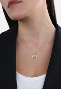 Collana d'argento con pendente a simbolo dell'infinito, circondata da piccoli accenti rotondi, poggiata su una camicia bianca sotto un blazer nero.