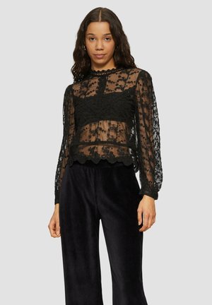 Zwarte doorschijnende blouse met bloemenborduursel, lange mouwen, gegolfde zoom en een hoge halslijn. Het model draagt het boven donkere, wijde broeken.