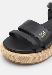 Black leather sandal featuring two wide straps, a woven jute platform sole, and a metallic logo accent on the upper.
