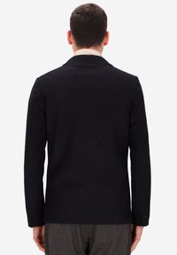 Un uomo in giacca nera e pantaloni grigi si trova di spalle contro uno sfondo bianco uniforme.