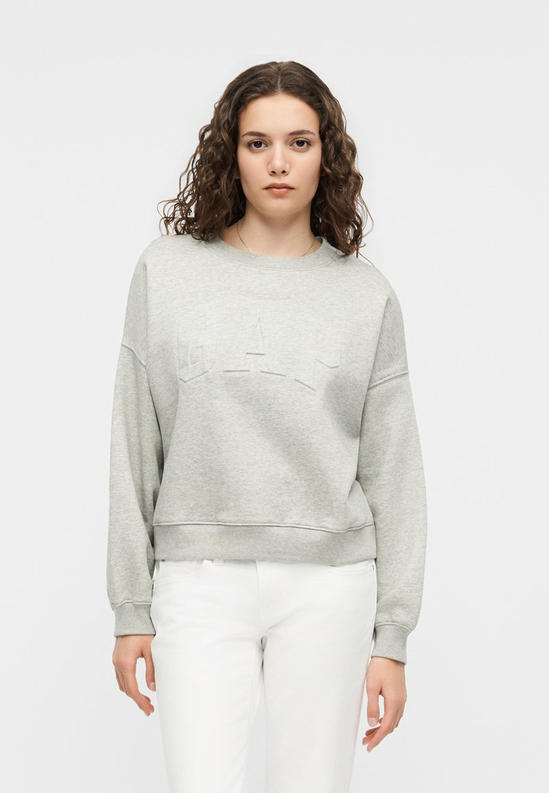 Νεαρή γυναίκα με σγουρά μαλλιά που φοράει ανοιχτό γκρι φούτερ και λευκό παντελόνι, στέκεται μπροστά σε ένα απλό λευκό φόντο.