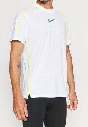 Mężczyzna w białej sportowej koszulce Nike z neonowożółtymi szwami i czarnych spodniach, stojący na jednolitym tle.