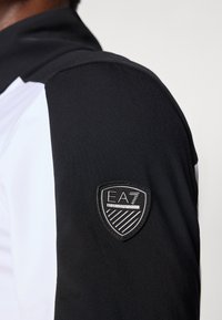 Mustvalge sportlik jakk, millel on kõrge krae, sile kangas ja EA7 logo märk vasakul õlal.