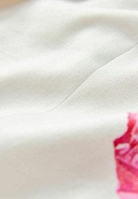 Baumwollstoff mit einer glatten Textur, hauptsächlich weiß mit einem rosafarbenen Blumenmuster an einer Kante, das ein leichtes und sanftes Erscheinungsbild hervorhebt.