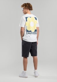 Valkoinen puuvillapaita, jossa on suuri keltainen kukkakuvasto ja "CALIFORNIA" -teksti selässä, yhdistettynä tummiin shortseihin ja valkoisiin lenkkareihin.