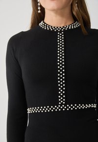 Fekete kötött ruha, kerek kivágással, amelyet gyöngyszerű díszítések sorai ékesítenek, plusz részletek a deréknál és az elülső középen.