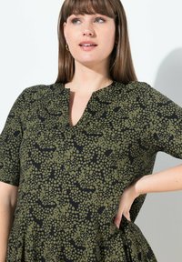 Vestido de estampado verde y negro con escote en V, mangas cortas y bolsillos laterales; presenta una tela suave y texturizada, y un corte relajado.