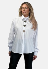 Biała koszula z guzikami, z wyraźnym kołnierzykiem, trzema dużymi srebrnymi guzikami na przodzie oraz długimi, bufkowymi rękawami. Gładka faktura materiału.