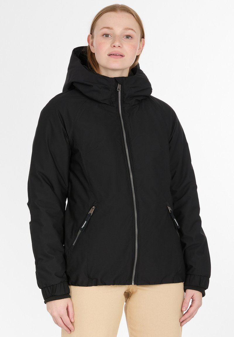 Donna che indossa una giacca a zip nera con cappuccio e tasche laterali con zip, con pantaloni beige, in piedi davanti a uno sfondo bianco.