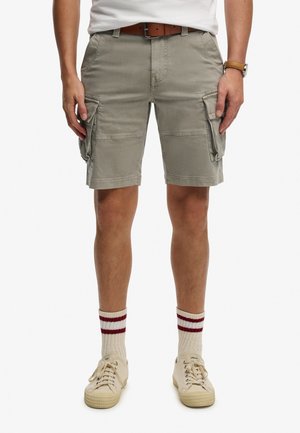 Man draagt lichtgrijze cargoshorts, witte sneakers, witte sokken met rode strepen, bruine riem en wit T-shirt, staat met ontspannen handen.