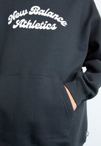 Hoodie noir avec le texte blanc "New Balance Athletics" sur la poitrine et la main partiellement dans la poche.