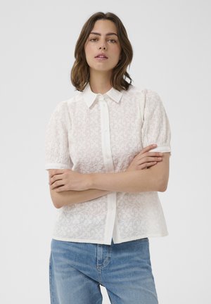 Noor naine, kellel on õlgadeni pruuni juuksed, kandes valget lühikeste varrukatega pitspluusi ja siniseid teksaseid, käed risti.