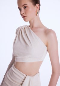Top cropped asymétrique à une épaule en beige clair, avec un tissu drapé et une texture lisse. Associé à une jupe assortie.