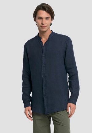 Giovane uomo che indossa una camicia blu scuro con bottoni e collo alla coreana e pantaloni verde oliva, in piedi davanti a uno sfondo chiaro e uniforme.