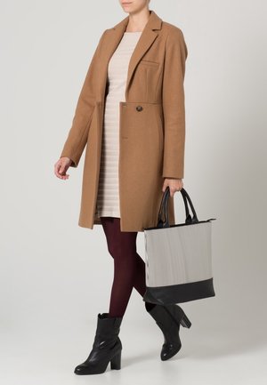 Manteau en laine camel avec une coupe cintrée, robe beige rayée, collants bordeaux, bottines noires et sac fourre-tout rayé avec des accents noirs.