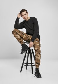 Čierna košeľa s dlhým rukávom, maskárové jogger nohavice s elastickými manžetami a čierne vysoké tenisky. Model sedí na čiernej stoličke.